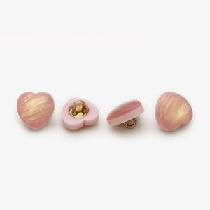 Peut inclure: Quatre boutons en forme de cœur de couleur rose clair avec un motif tourbillonnant doré. Chaque bouton est doté d'une tige dorée pour la couture. Les boutons sont probablement destinés à des vêtements ou à des projets d'artisanat.
