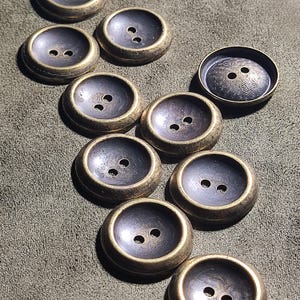 May include: A collection of round, two-hole buttons featuring a dark, recessed centre and a metallic, antique-gold rim. One button displays a distinct circular pattern and the text 21.25 engraved on its surface. The buttons have a smooth, concave design.