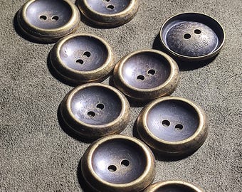 May include: A collection of round, two-hole buttons featuring a dark, recessed centre and a metallic, antique-gold rim. One button displays a distinct circular pattern and the text 21.25 engraved on its surface. The buttons have a smooth, concave design.