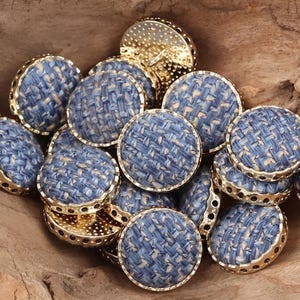 May include: A collection of round, blue and cream fabric covered buttons with gold metal trim. The buttons are arranged in a pile on a wooden surface.