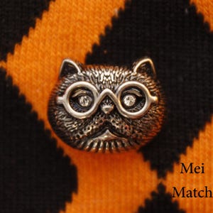 May include: A silver-toned cat figurine with round glasses and rhinestones for eyes. The cat is wearing a serious expression.