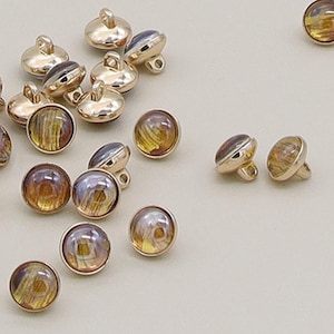 Peut inclure: Plusieurs boutons à tige dorés avec un centre en forme de dôme, irisé et semblable à du verre. Les boutons ont un bord doré et une boucle pour la couture. Les centres affichent des nuances d'ambre, de marron et de violet.