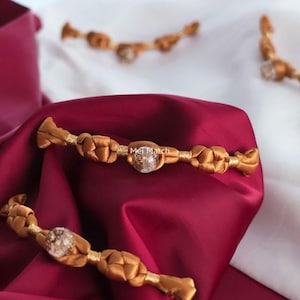 May include: Gold-colored hair accessories with a braided design and a clear, round gem in the center. The accessories are displayed on a burgundy and white satin background. The text "Mei Match" is visible.