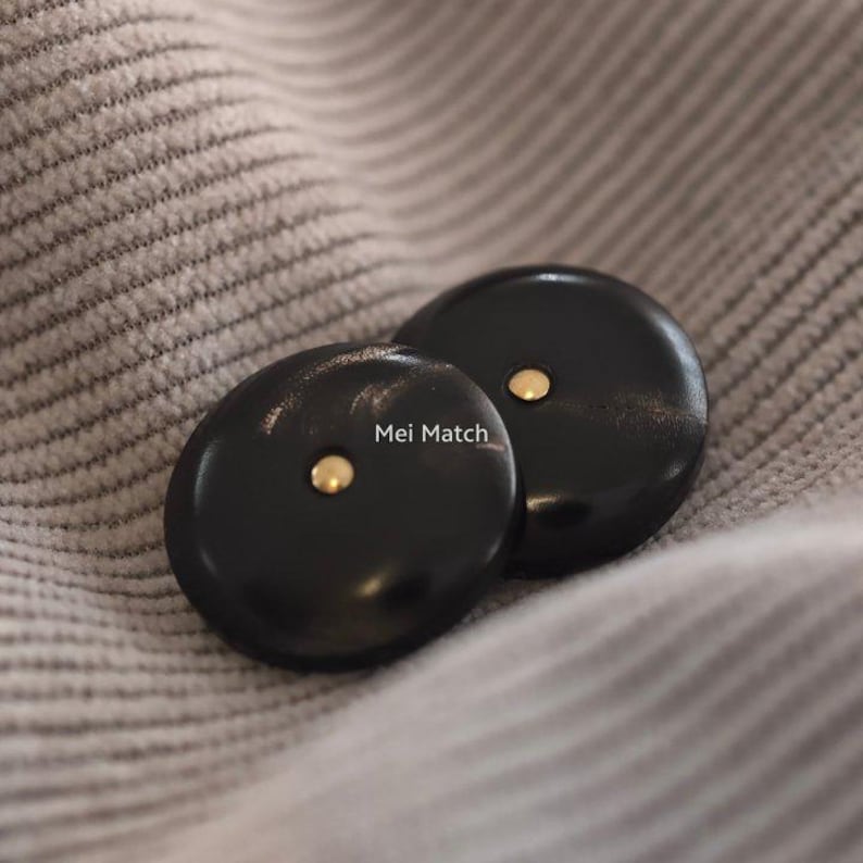 May include: Two round black buttons with a smooth, polished finish and a small, centered gold-tone metal stud. The text Mei Match is printed in white between the two buttons. The buttons have a slightly domed shape and a glossy surface texture.