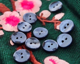 May include: A collection of ten round, dark navy blue buttons, each featuring two central holes. The buttons are scattered across a dark green fabric surface decorated with embroidered pink flowers and brown branches. The text Mei Match is visible.