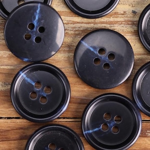 Può includere: Una collezione di bottoni neri con un design marmoreo blu e quattro fori ciascuno. I bottoni sono disposti su una superficie di legno.