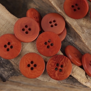 Puede incluir: Una colección de 10 botones de madera marrón, cada uno con cuatro agujeros. Los botones están dispuestos en un semicírculo sobre una superficie de madera.