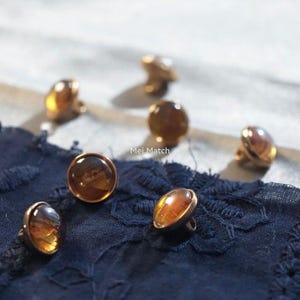 Peut inclure: Gros plan sur plusieurs boutons ambrés avec des bordures en métal doré. Les boutons sont disposés sur un tissu bleu foncé, avec le texte "Mei Match" visible en arrière-plan. Les boutons ont un aspect bombé et translucide.