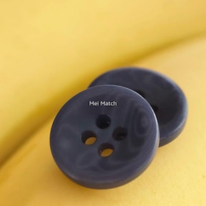 May include: Two round, dark charcoal gray buttons featuring a subtle marbled pattern and four central holes for attachment. The text Mei Match is printed in white on the surface of the front button, which has a smooth, slightly concave center.