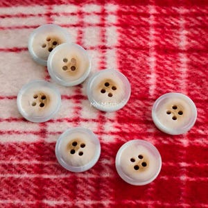 Puede incluir: Un conjunto de siete botones redondos con cuatro agujeros cada uno. Los botones tienen un anillo exterior azul claro y un centro color crema. El texto "Mei Match" es visible en un botón. Los botones están dispersos sobre una tela a cuadros roja y blanca.