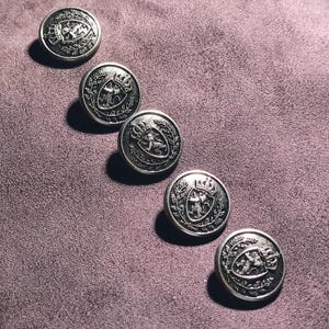 Peut inclure: Cinq boutons métalliques circulaires de couleur argentée disposés en ligne diagonale. Chaque bouton présente un motif en relief représentant un bouclier surmonté d'une couronne, contenant un lion, entouré d'une bordure décorative de feuilles.