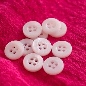 May include: A collection of round, light pink buttons, each featuring four central holes for attachment. The buttons have a smooth, slightly glossy finish and are arranged in a cluster. The text Mei Match is visible on one button.