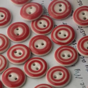 May include: A selection of red and white striped buttons, each with two holes. The buttons are circular, featuring a white centre and a red and white striped outer ring. They are arranged on a white surface.