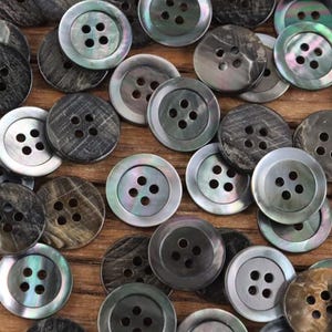 May include: A selection of round buttons in shades of grey, black, and iridescent hues. Each button features four holes and a subtly raised edge. The buttons are arranged on a wooden surface, creating a textured and visually appealing display.