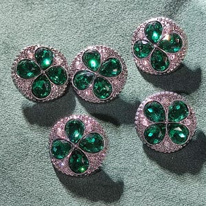 Pode incluir: Cinco botões redondos prateados com um design de trevo. Cada botão apresenta quatro pedras verdes em forma de gota, rodeadas por cristais transparentes menores. Os botões estão dispostos sobre uma superfície verde.
