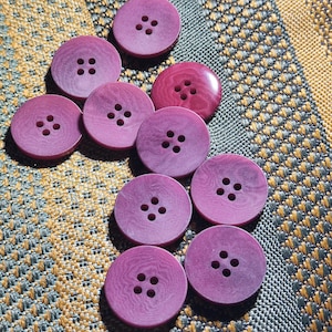 Puede incluir: Un conjunto de diez botones redondos de color morado, cada uno con cuatro orificios centrales para su fijación. Los botones muestran una sutil textura de vetas de madera en sus superficies, con un botón que parece más brillante que los demás.