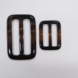Könnte beinhalten: Zwei glänzende, rechteckige Schnallen mit einem marmorierten braun-schwarzen Muster. Die größere Schnalle hat zwei vertikale Schlitze und abgerundete Ecken. Die kleinere Schnalle spiegelt das Design wider und bietet ein passendes Set.