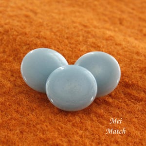 Puede incluir: Tres botones redondos de color azul claro con un acabado ligeramente brillante. Los botones están sobre una superficie naranja peluda.