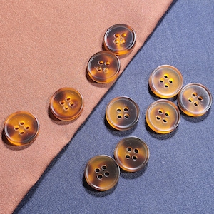 Può includere: Una collezione di dieci bottoni rotondi a quattro fori con un motivo tartarugato traslucido color ambra e marrone. I bottoni sono sparsi su una superficie divisa diagonalmente tra un tessuto testurizzato color terracotta e un tessuto blu scuro.