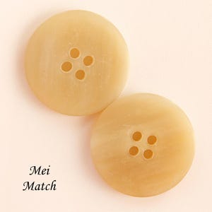May include: Two large, round, light tan buttons with four holes each. The buttons have a slightly marbled appearance and are set against a soft, off-white background. The text "Mei Match" is visible in the lower left corner.