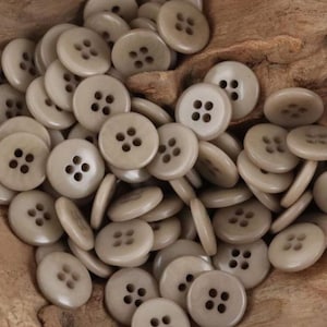 May include: A pile of beige, round buttons with four holes each. The buttons are scattered, some lying flat and others on their sides, creating a textured, tactile appearance. The buttons are made of a smooth material.