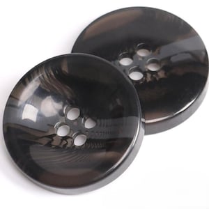 Puede incluir: Dos botones grandes y redondos de color gris oscuro con cuatro agujeros cada uno. Los botones tienen un acabado brillante y un sutil patrón arremolinado. Los botones son para proyectos de costura o manualidades.