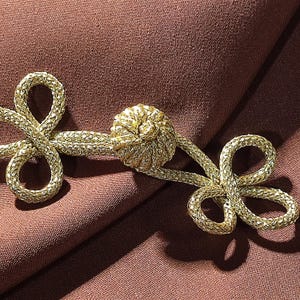 May include: A close-up shot of a gold frog closure. The closure has a central knot and two looped ends, all made from a woven material. The background is a plain brown fabric.