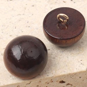 Puede incluir: Dos botones de madera marrón oscuro con acabado brillante. Un botón tiene un lazo metálico dorado en la parte superior para coser. Los botones son redondos y tienen un aspecto liso y pulido. Ideal para proyectos de costura.