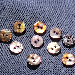 Puede incluir: Una colección de botones redondos e iridiscentes, cada uno con dos agujeros. Los botones muestran una gama de colores, incluyendo dorado, marrón y rosa, con un brillo nacarado. Los botones están sobre una tela azul oscuro.