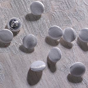 Könnte beinhalten: Eine Ansammlung von zehn silbernen, stoffbezogenen Knöpfen, die auf einer strukturierten Oberfläche verstreut sind. Die Knöpfe sind rund mit leicht gewölbter Form und glänzendem Metallrücken. Der Stoff hat ein dezentes Webmuster.