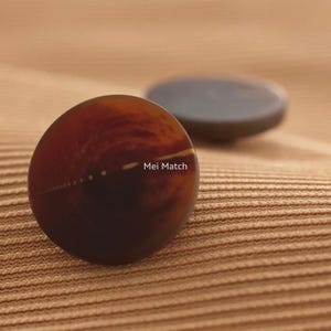 May include: Two round, dark brown buttons with a smooth, polished finish and a subtle marbled pattern. The front button features the text Mei Match printed in white. The buttons have a circular shape and a slightly domed surface profile.