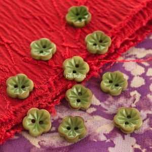 May include: Olive green flower-shaped buttons with two holes, scattered on red and purple patterned fabric. The buttons feature a detailed petal design. The text "Mei Match" is visible on the fabric, suggesting a brand or maker.