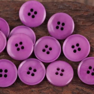 以下が含まれることがあります： 丸い紫色のボタンのコレクション。各ボタンには4つの黒い穴があります。ボタンは暗く、質感のある表面に配置されています。これらのボタンは、縫製や手芸プロジェクトに適した、光沢のある仕上がりの単色です。