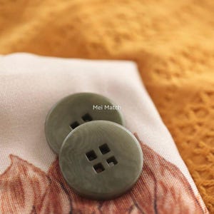 May include: Two round, olive green buttons with a subtle marbled texture and four square holes in the centre. The text Mei Match is printed in white above the buttons. The buttons have a smooth, matte finish and a flat, circular design.
