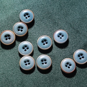 Può includere: Una collezione di dieci bottoni azzurri con quattro fori ciascuno. I bottoni hanno un bordo color legno chiaro e sono disposti su una superficie verde scuro. Questi bottoni sono adatti per progetti di cucito e artigianato.