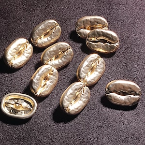 May include: A collection of metallic gold-toned buttons shaped like coffee beans. Each button features a textured, uneven surface with a central crevice. One button is flipped over to reveal a flat back with a central shank and the text ZPHE 20.