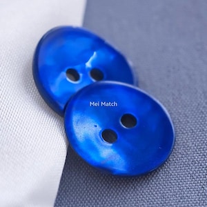 May include: Two round, vibrant blue buttons with a smooth, glossy finish and two central holes each. The text Mei Match is printed in white on the surface of the bottom button. These circular fasteners feature a slightly irregular, organic edge.