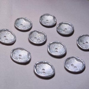 Pode incluir: Um conjunto de nove botões circulares de tom prateado, cada um apresentando uma borda irregular e orgânica, e um padrão texturizado de anéis concêntricos na superfície. Cada botão possui dois orifícios centrais para fixação.
