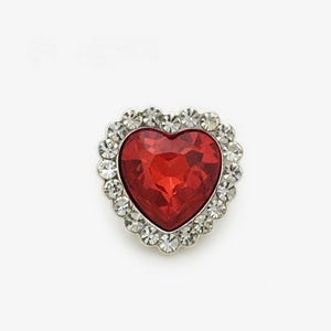 Puede incluir: Una joya roja en forma de corazón con un borde plateado adornado con pedrería. La joya es de color rojo intenso y tiene una superficie facetada. Los brillantes son claros y redondos, creando un efecto brillante. Probablemente un broche o colgante.
