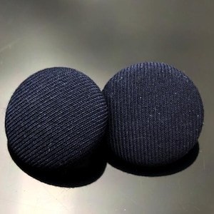 Può includere: Due bottoni rotondi blu navy rivestiti in tessuto. I bottoni hanno una trama a coste e sono posizionati uno accanto all'altro. Questi bottoni sono probabilmente per abbigliamento o progetti artigianali.