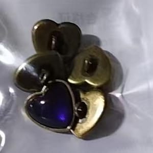 May include: A collection of heart-shaped charms in various metallic finishes. One charm features a deep blue, possibly glass, heart set within a silver-toned frame. The other charms have a gold-toned finish.
