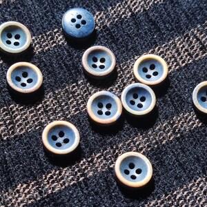 May include: A collection of ten round, four-hole buttons featuring a dark blue center and a light tan outer rim. Each button has a recessed circular design with four evenly spaced holes in the middle, arranged in a scattered pattern.
