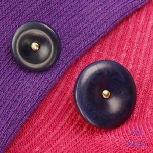 May include: Two dark blue round buttons with a gold center on a purple and pink fabric background.