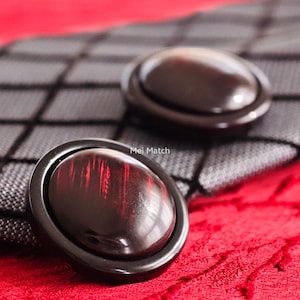 Puede incluir: Dos botones ovalados de color marrón oscuro con un acabado brillante y un borde negro. Los botones tienen un patrón de vetas de madera marrón rojizo. Están sobre una tela a cuadros negra y gris, con una tela roja en el fondo. El texto "Mei Match" es visible.