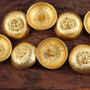 Puede incluir: Primer plano de varios botones dorados con un diseño texturizado y acanalado. Cada botón tiene un adorno floral central y un ojal para coser. Los botones están dispuestos sobre una superficie de madera marrón oscuro.