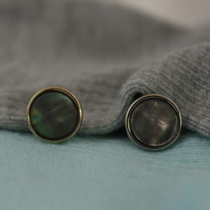 Puede incluir: Dos botones redondos decorativos con un acabado iridiscente negro y verde. Un botón tiene un borde dorado y el otro un borde plateado.