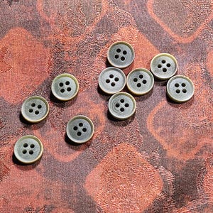 May include: A collection of nine round, four-hole buttons featuring a dark gray center and a metallic gold-toned rim. Each button has a smooth, circular design with four evenly spaced holes in the middle, arranged in a square pattern for attachment.