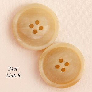 May include: Two large, round, light tan buttons with four holes each. The buttons have a slightly marbled appearance and are set against a pale pink background. The text "Mei Match" is in the lower left corner.