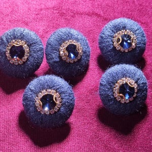 May include: Five navy blue fabric buttons with a central blue jewel surrounded by a ring of clear rhinestones and a gold-colored metal frame. The buttons are arranged on a pink surface.