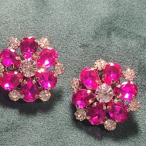 May include: A pair of circular earrings featuring a central clear round rhinestone surrounded by six teardrop-shaped hot pink rhinestones and six smaller clear round rhinestones arranged in a floral pattern with gold-toned metal settings.